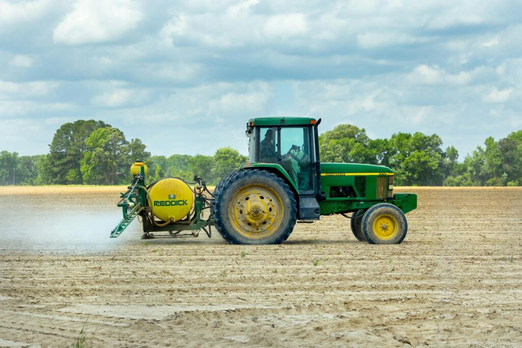 pexels-photo-2889440-2889440 Green tractor with sprayer working in an open farm field under a cloudy sky.